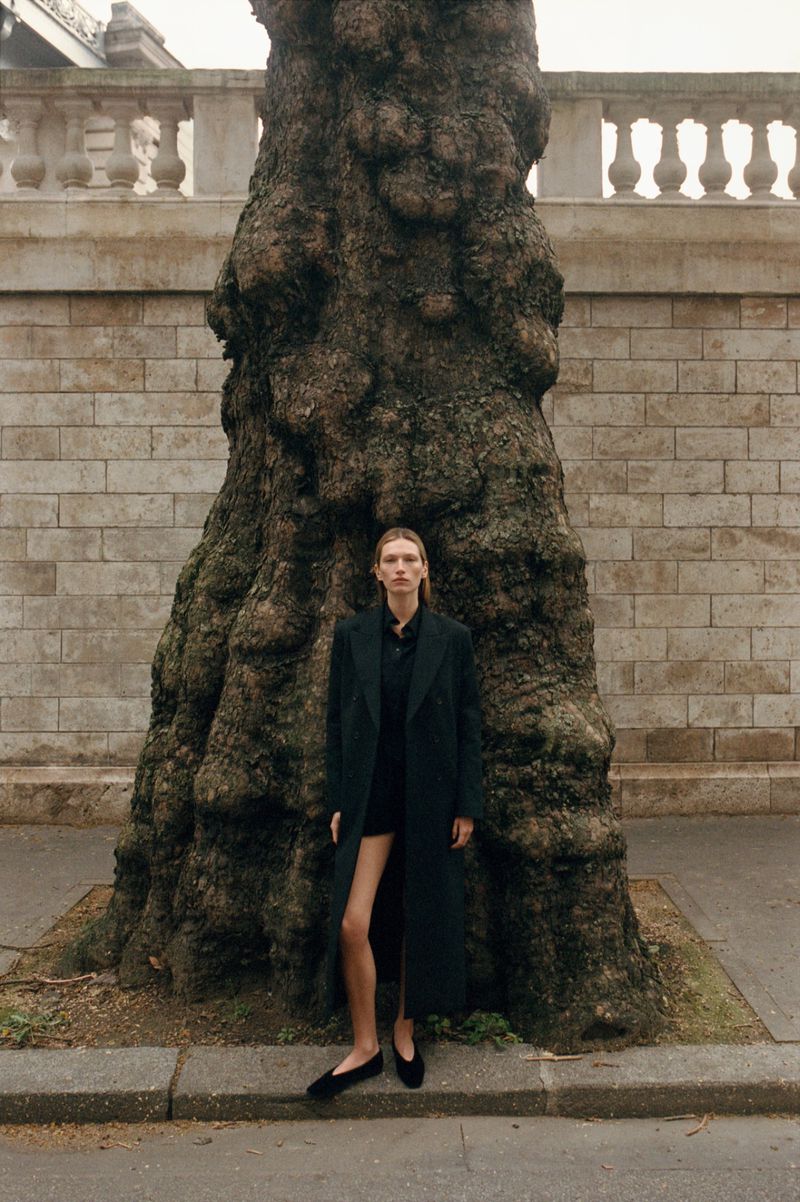 Model standing in front of a massive textured tree trunk wearing a long dark coat and flats against a stone wall background.