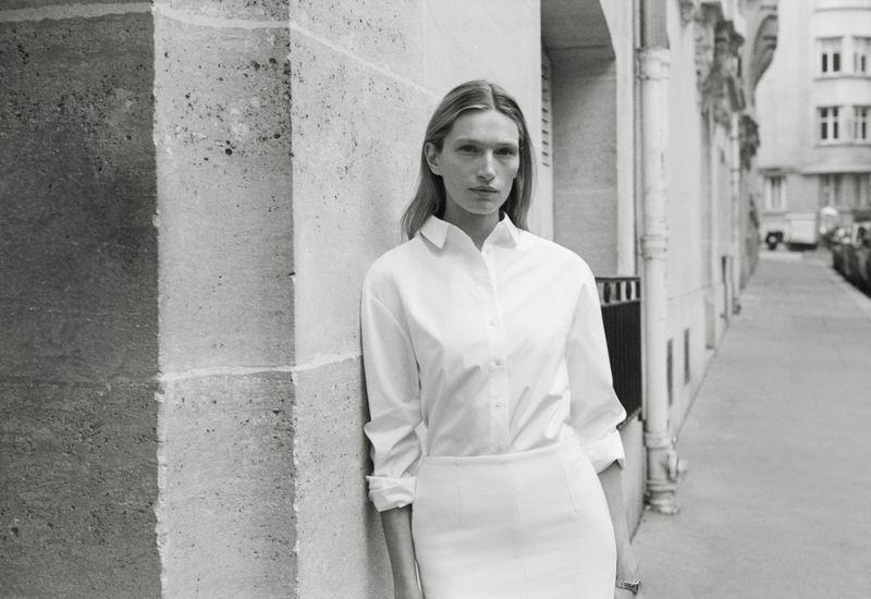 Model Liz Kennedy leans against a stone building wearing a white button-down shirt for the Almada S/S 2026 campaign.