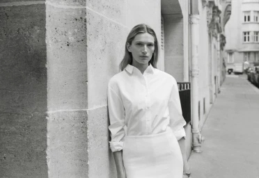 Model Liz Kennedy leans against a stone building wearing a white button-down shirt for the Almada S/S 2026 campaign.