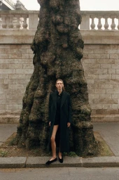 Model standing in front of a massive textured tree trunk wearing a long dark coat and flats against a stone wall background.