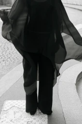 Model in flowing black sheer cape and wide trousers standing on a paved street