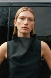 Close-up portrait of model Liz Kennedy wearing a black sleeveless top against a dark vehicle exterior for ALMADA S/S 2026.