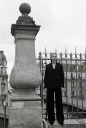 Model standing outdoors wearing a dark suit jacket and trousers near an ornate stone pillar and metal fence