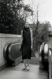 Monochrome fashion portrait of a model standing at the top of an outdoor escalator wearing a dark top and skirt holding a clutch.