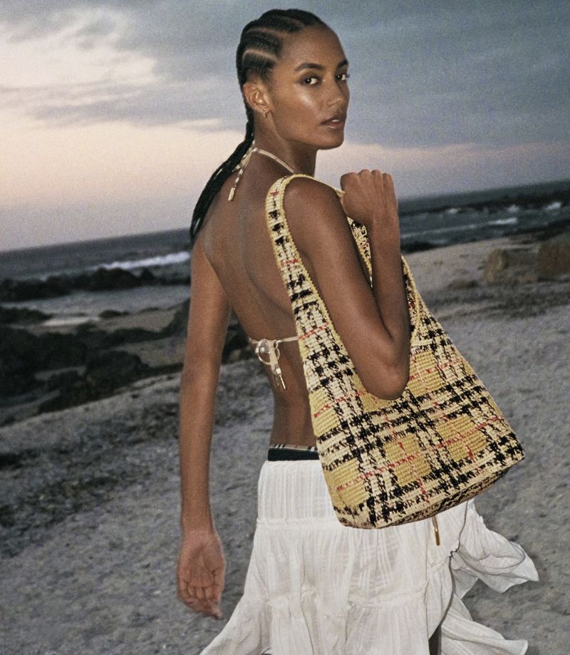 Model standing on a sandy beach at dusk holding a woven plaid tote bag for the Burberry High Summer 2026 campaign.