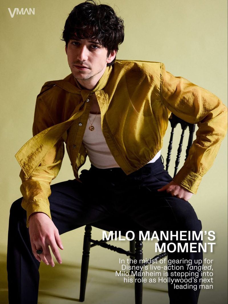Milo Manheim sitting on a black chair wearing a sheer gold button-down shirt and black trousers against a pale olive backdrop