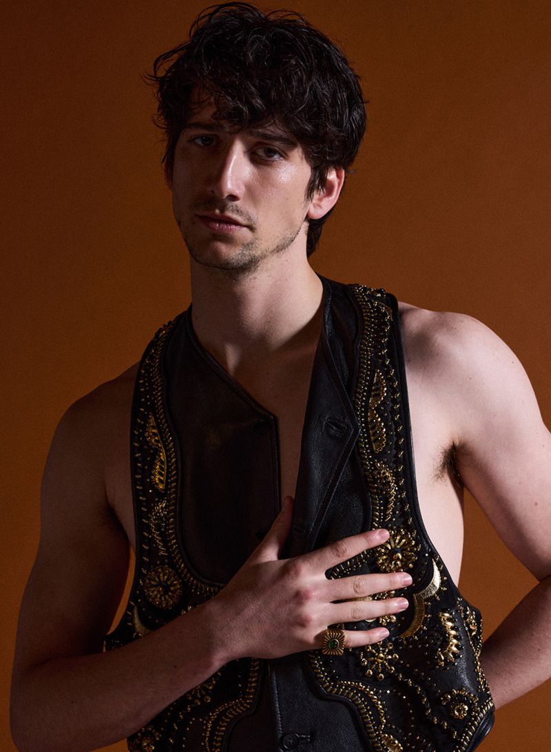 Portrait of a man wearing a black leather vest with ornate gold embroidery against a solid brown background