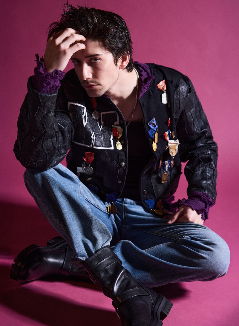 Milo Manheim sitting cross-legged wearing a decorated black jacket and blue jeans against a magenta background for V Man.