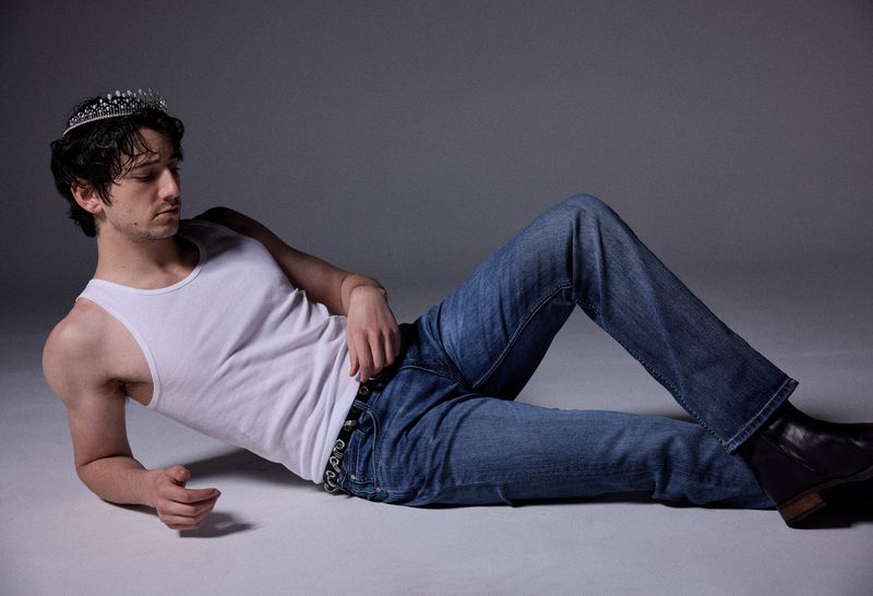 Milo Manheim wearing a white tank top, blue jeans, and a tiara while reclining on a grey studio floor for V Man.