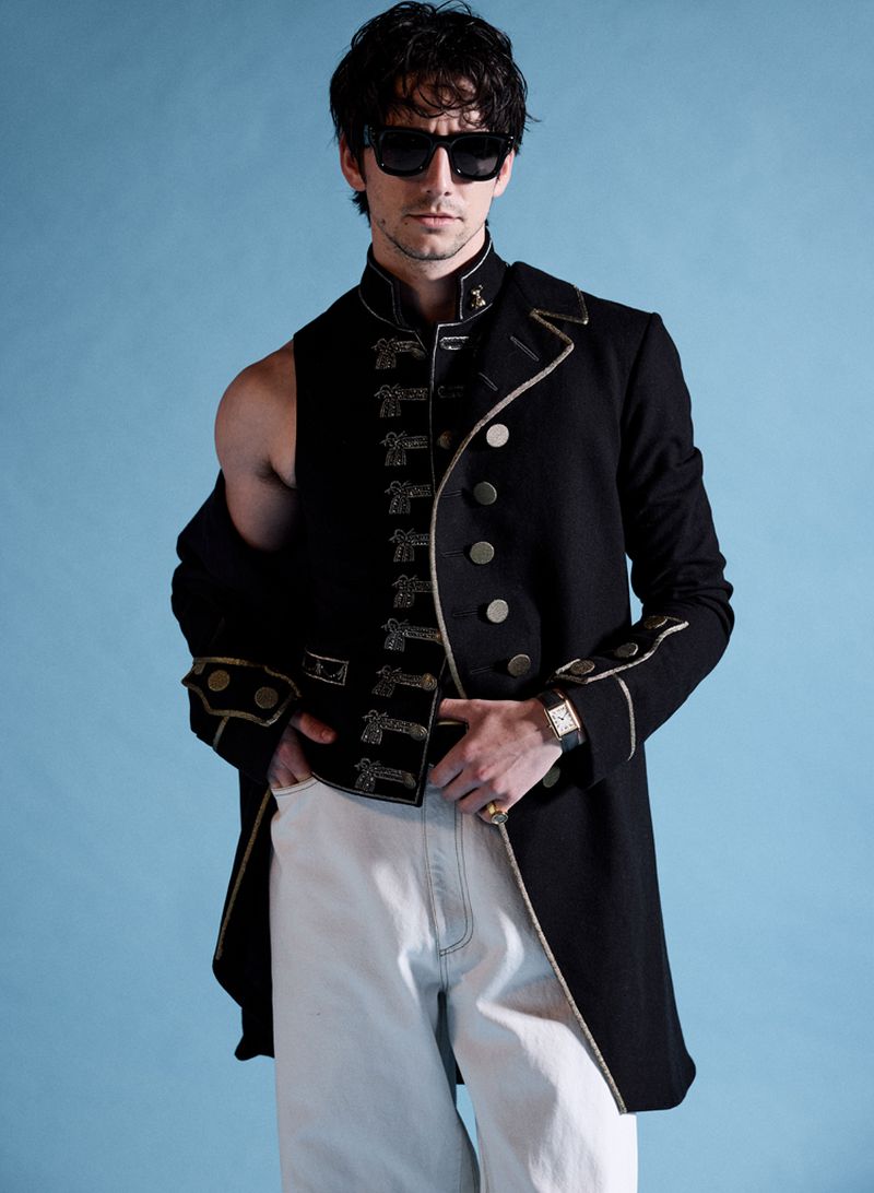 Adult male in sunglasses wearing a black coat with gold trim and white denim pants against a light blue background, indoor setting.