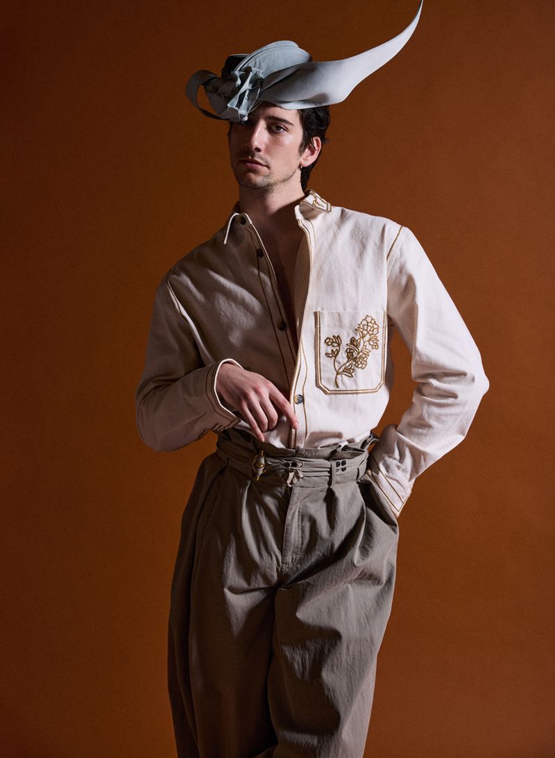 Man wearing a cream button-down shirt, grey pleated trousers, and a sculptural curved hat against a brown background.