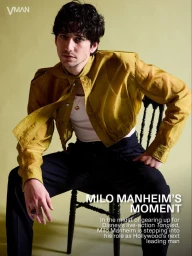 Milo Manheim sitting on a black chair wearing a sheer gold button-down shirt and black trousers against a pale olive backdrop