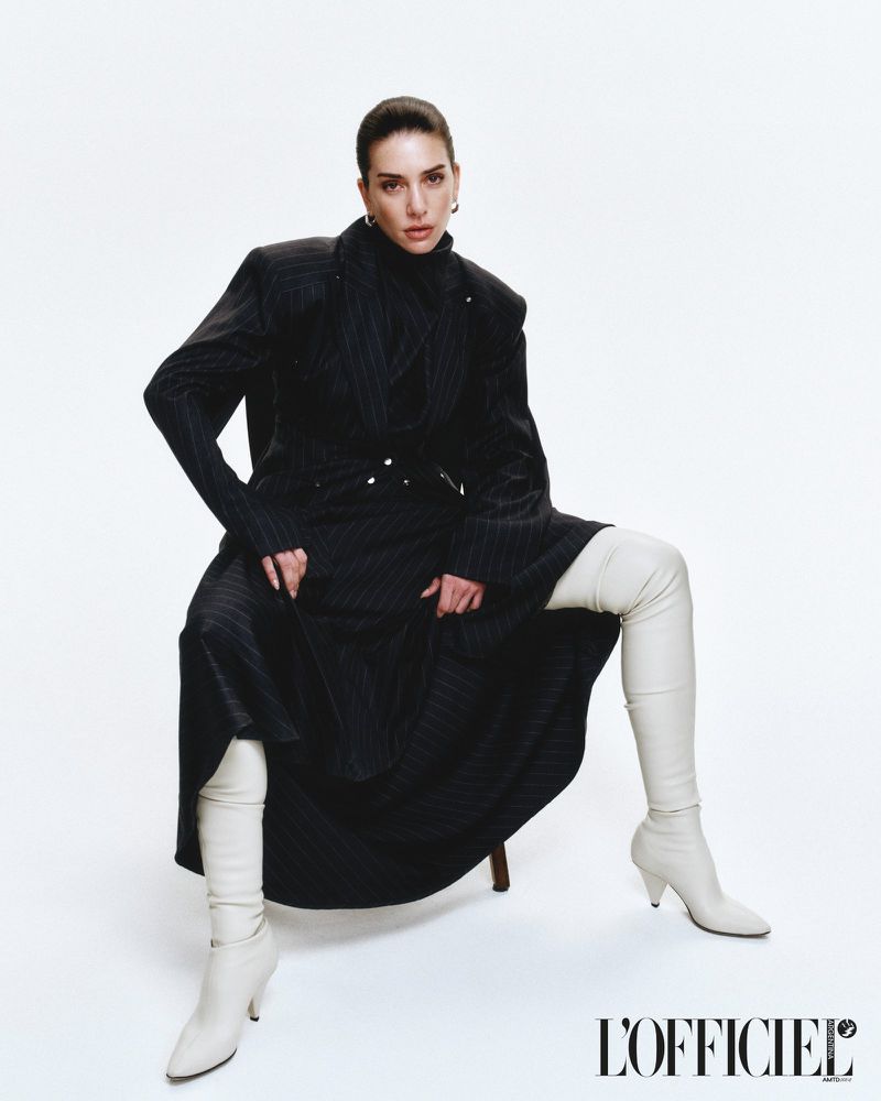 Fashion shot of a woman in a black coat and white thigh-high boots sitting on a stool against a white backdrop in a studio setting.