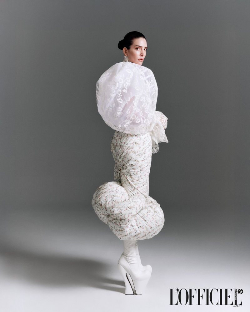 Model wearing sculptural white blouse and patterned skirt with unique silhouette against grey studio backdrop