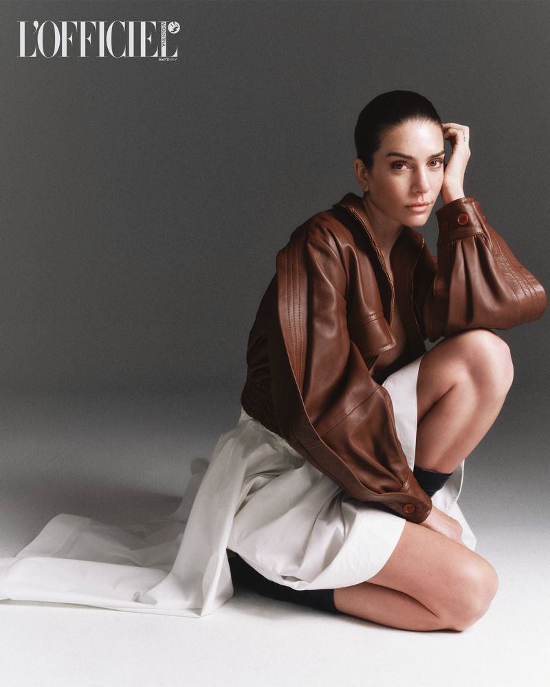 Model in brown leather jacket and white skirt crouching against a dark grey studio background