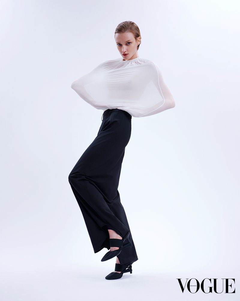 Fashionable adult woman poses in black pants and a draped white top against a white backdrop in a studio, showcasing high fashion.