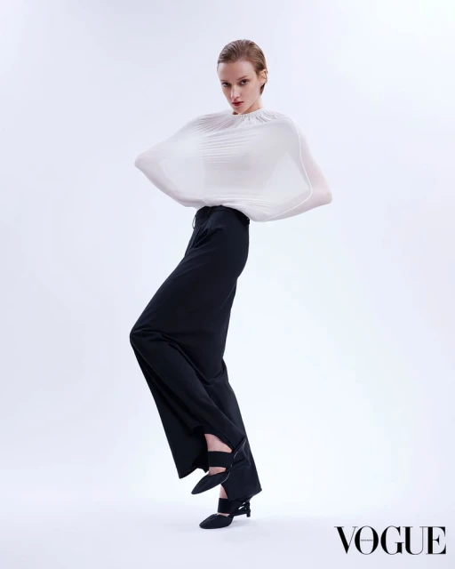 Fashionable adult woman poses in black pants and a draped white top against a white backdrop in a studio, showcasing high fashion.