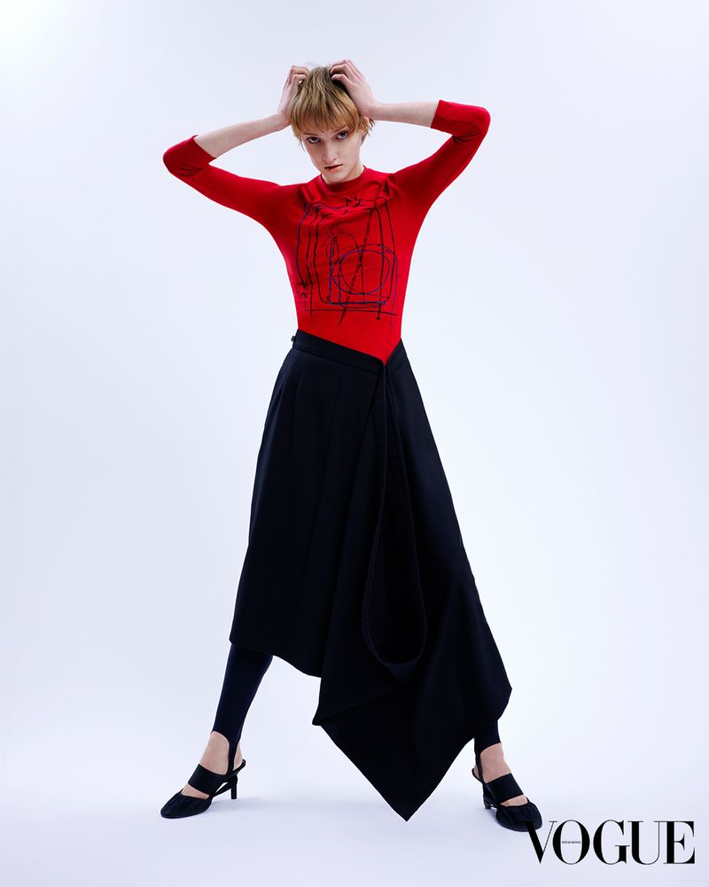 A young adult fashion model poses in a red top and black skirt against a white backdrop, holding her hands on her head in a studio shot.