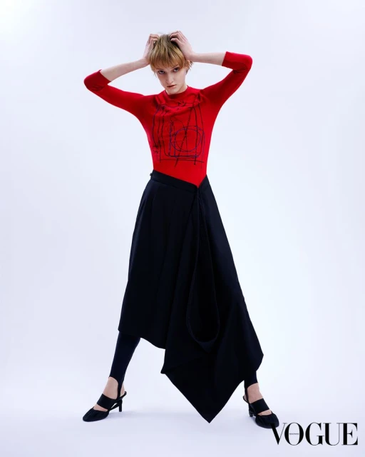 A young adult fashion model poses in a red top and black skirt against a white backdrop, holding her hands on her head in a studio shot.