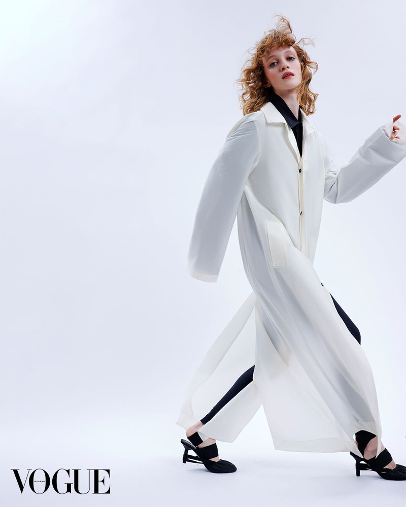 Fashion editorial shot of a redhead woman wearing a long white coat and black shirt, posing in a studio with a white background.