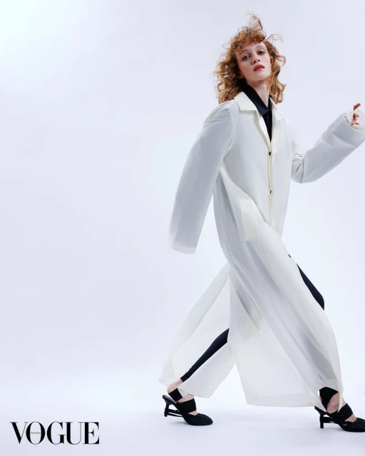 Fashion editorial shot of a redhead woman wearing a long white coat and black shirt, posing in a studio with a white background.