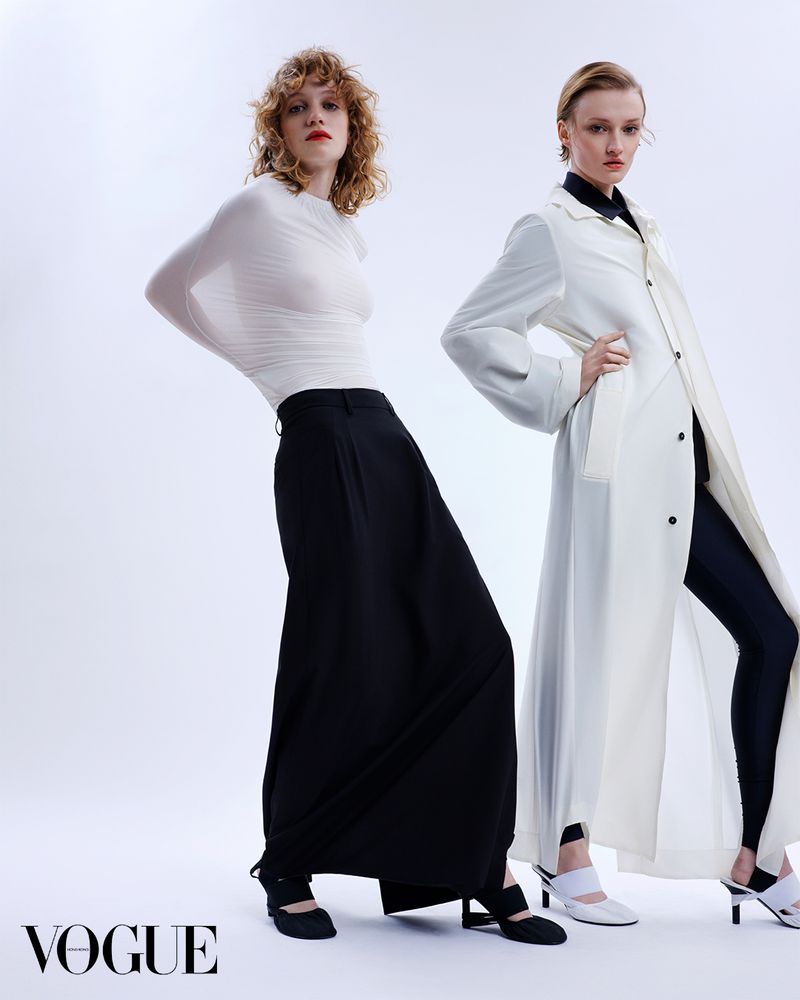 Two women model black and white fashion in a photo studio; one in a white top with a black skirt, the other in a white coat.