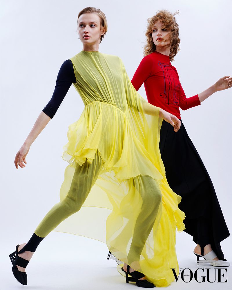 Two fashion models pose in studio wearing yellow and black contemporary couture and black strappy heels against a white backdrop.