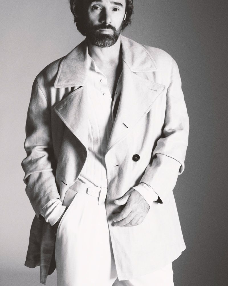 Monochrome portrait of an adult male with a beard wearing an open double-breasted coat, collared shirt and white pants in a studio.
