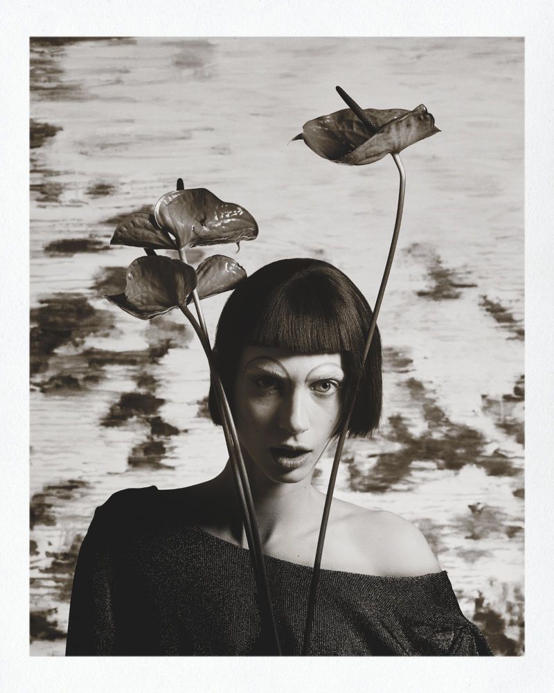 Black and white editorial portrait of a woman with short dark hair holding anthurium flowers for Marie Claire Belgium Beaton-Mania.