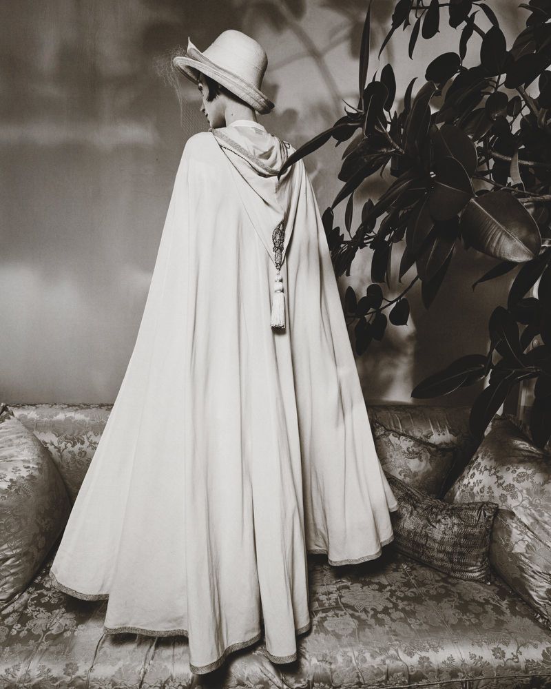 Model wearing a long white hooded cape and matching hat standing before a sofa and foliage in a monochromatic fashion editorial image