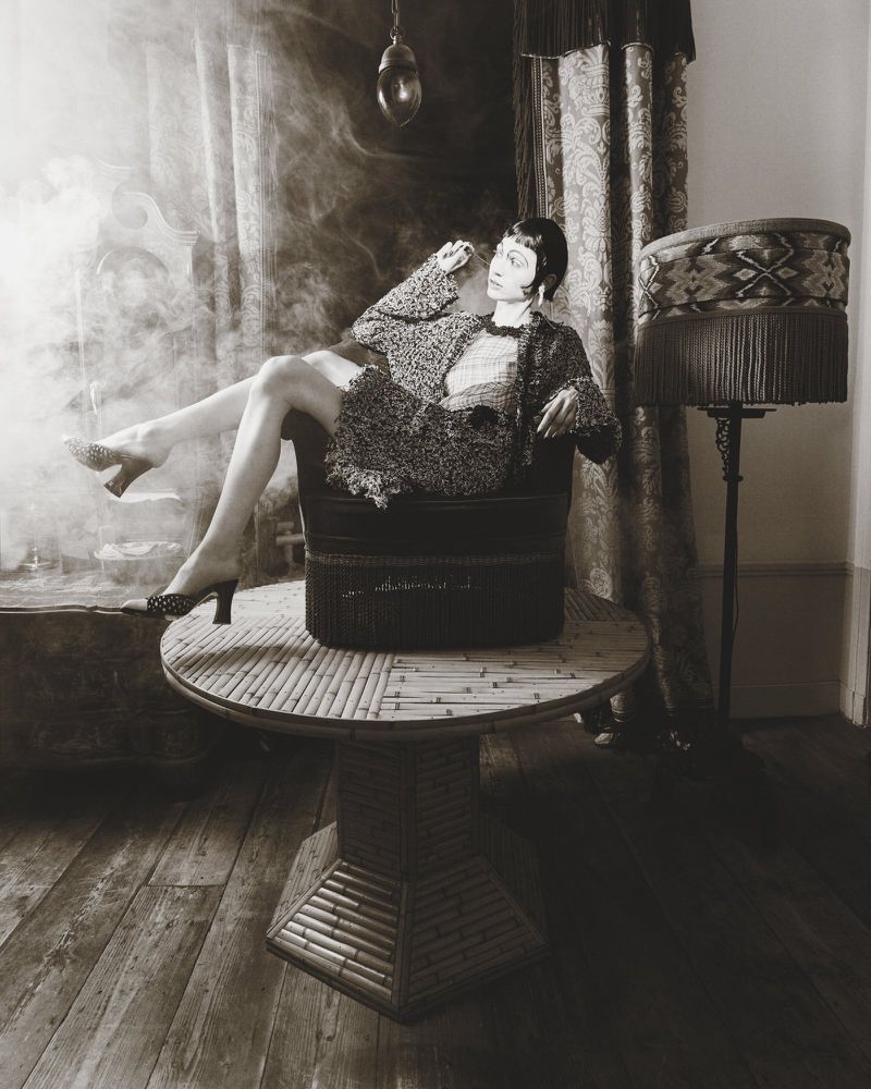 Woman in patterned coat seated in a chair on a table in a smoky, vintage-styled room for Marie Claire Belgium