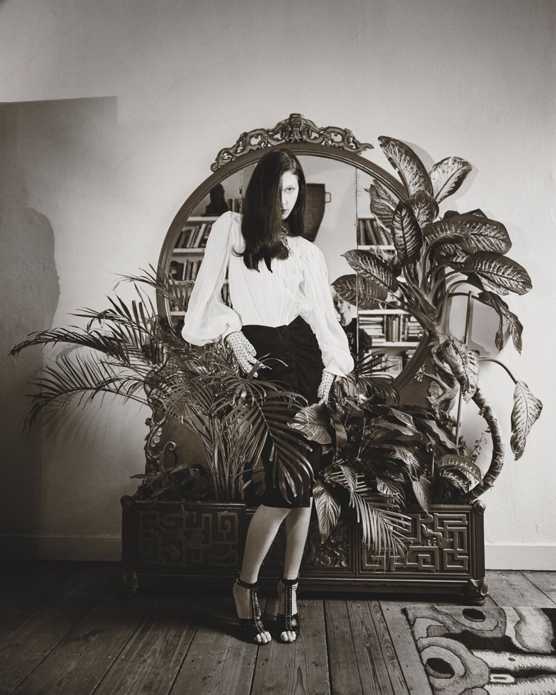 Model standing in front of an ornate mirror and potted plants wearing a white blouse and dark skirt for Marie Claire Belgium.