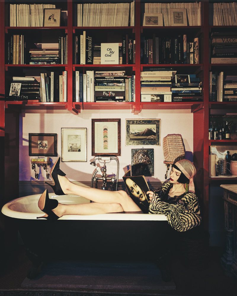 Woman in patterned top and headscarf reclining in a bathtub while reading a magazine, set against a red bookcase wall.
