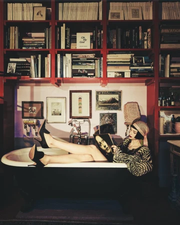 Woman in patterned top and headscarf reclining in a bathtub while reading a magazine, set against a red bookcase wall.