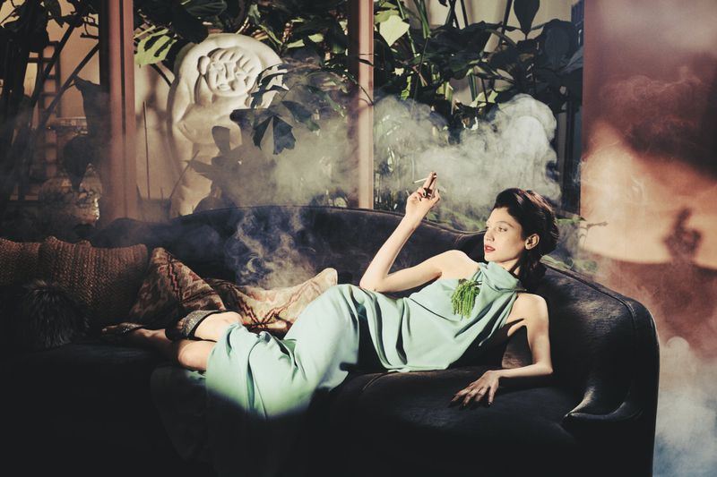 Woman in light green dress reclining on a dark velvet sofa holding a cigarette amidst wisps of smoke.