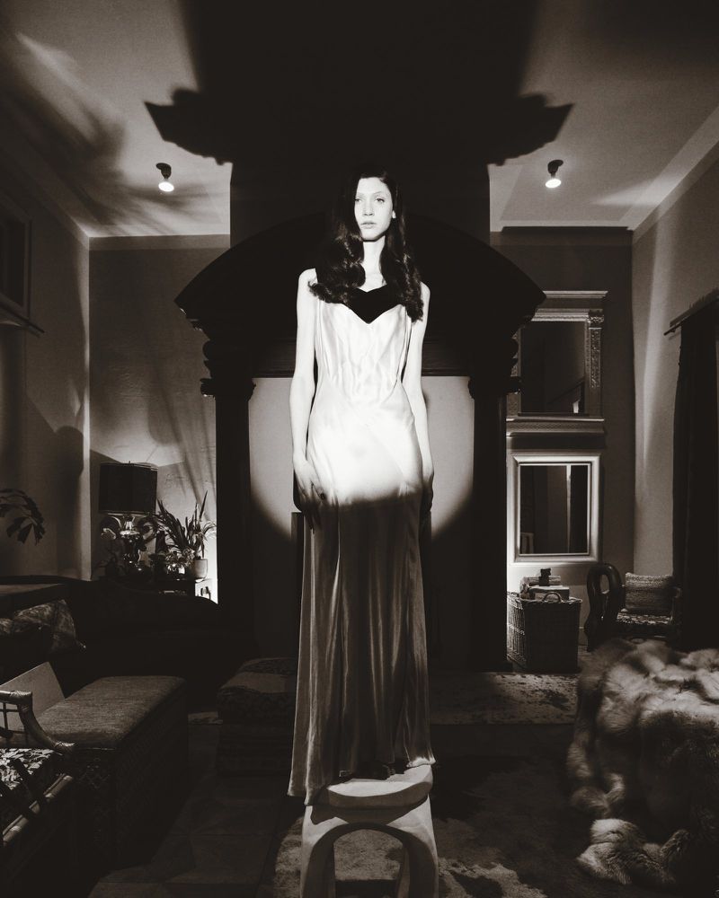 Model wearing a long evening gown standing on a small stool in a dimly lit living room for Marie Claire Belgium editorial shoot.