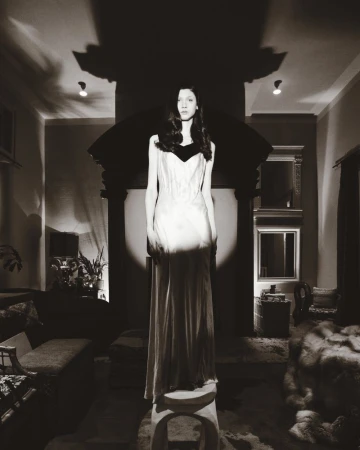 Model wearing a long evening gown standing on a small stool in a dimly lit living room for Marie Claire Belgium editorial shoot.