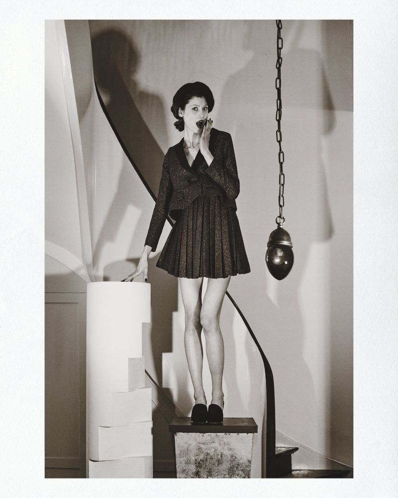 Monochrome fashion shot of a female model in a pleated dress and blazer, posing on a platform with architectural stairs and a hanging chain.