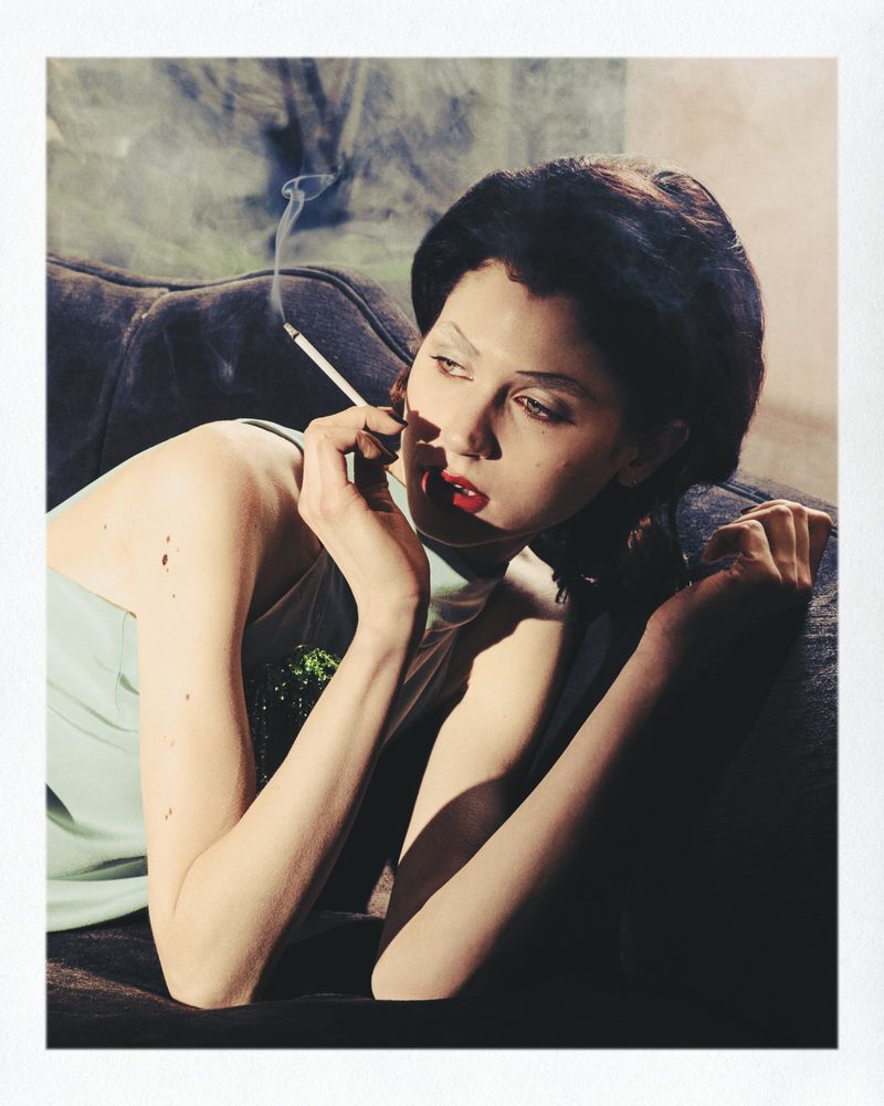 Woman in pale top lying on dark sofa holding a cigarette with smoke rising for Marie Claire Belgium Beaton-Mania editorial
