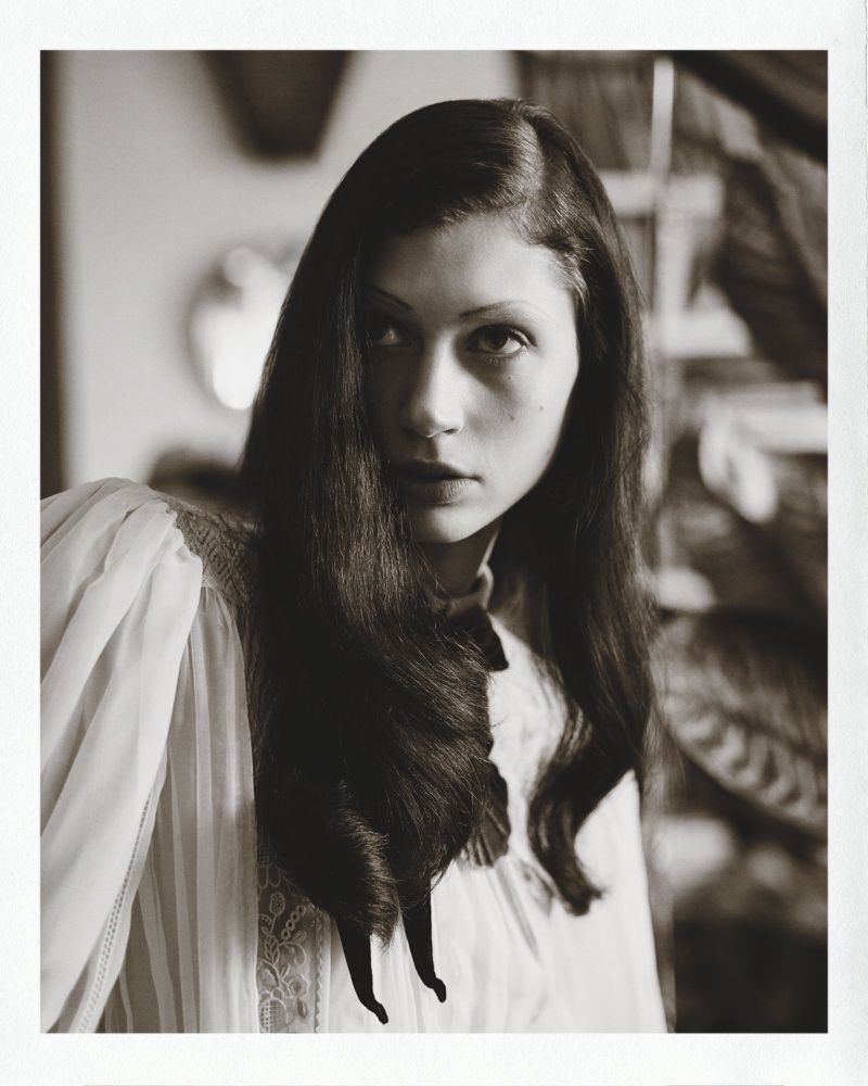 Portrait of a woman with dark wavy hair wearing a white pleated blouse with a dark bow, photographed for Marie Claire Belgium.