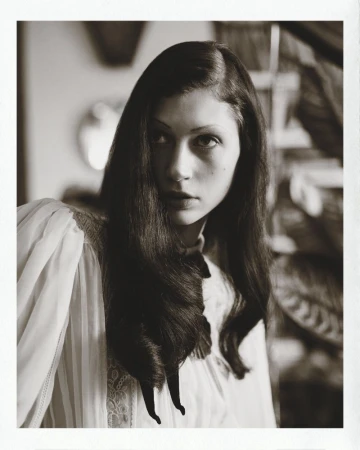 Portrait of a woman with dark wavy hair wearing a white pleated blouse with a dark bow, photographed for Marie Claire Belgium.
