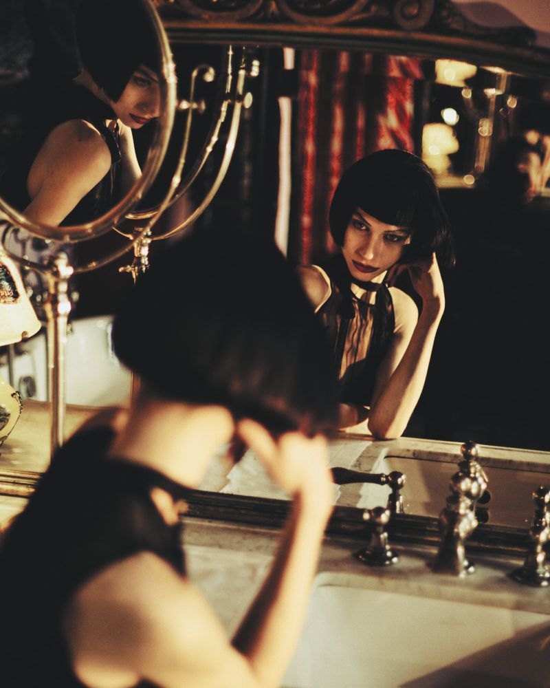 A dark-haired woman in a black top looking into a mirror in a dimly lit interior, photographed by Sieme Hermans for Marie Claire Belgium.