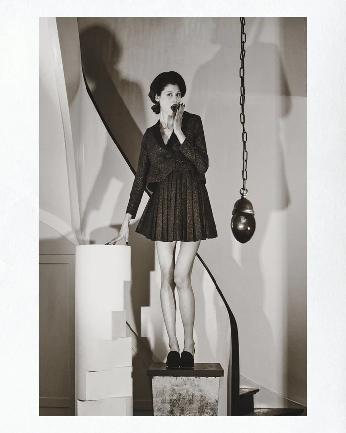 Model standing on a pedestal wearing a dark pleated skirt suit and cap with a hanging chain light in background