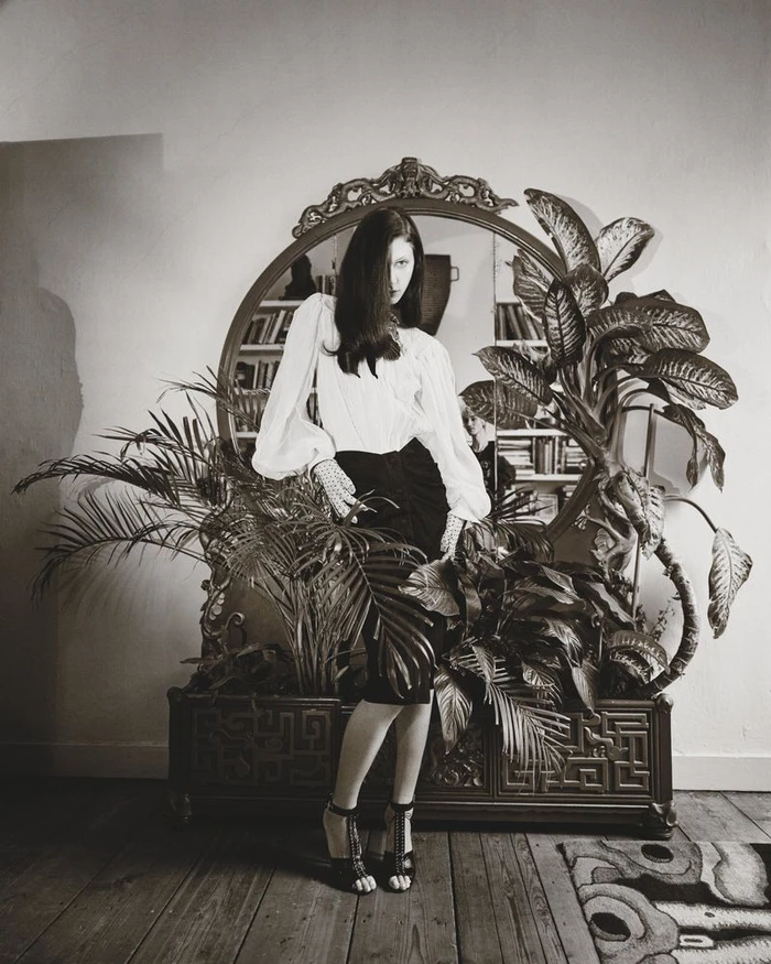 Model standing in front of an ornate mirror and potted plants wearing a white blouse and dark skirt for Marie Claire Belgium.