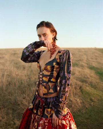 Model wearing patterned lace-up top and red skirt posing outdoors on a grassy hillside under a clear sky.