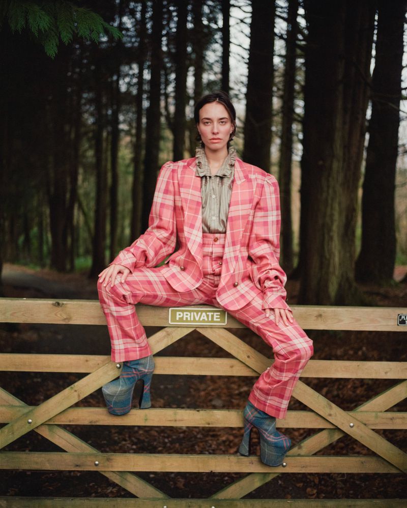 A woman in a pink plaid suit sits on a wooden gate in front of a forest. She wears platform shoes and a ruffled collar shirt.