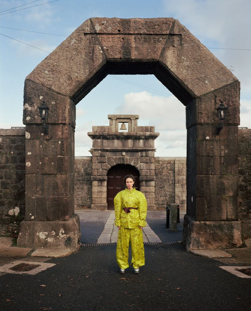 Model standing beneath a massive stone archway wearing a bright yellow patterned jacket and trousers with a brown belt.