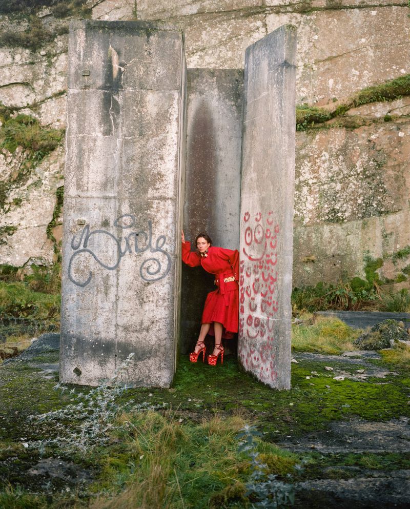 Model in a bright red coat and platform heels posing inside a weathered concrete structure on a rocky terrain