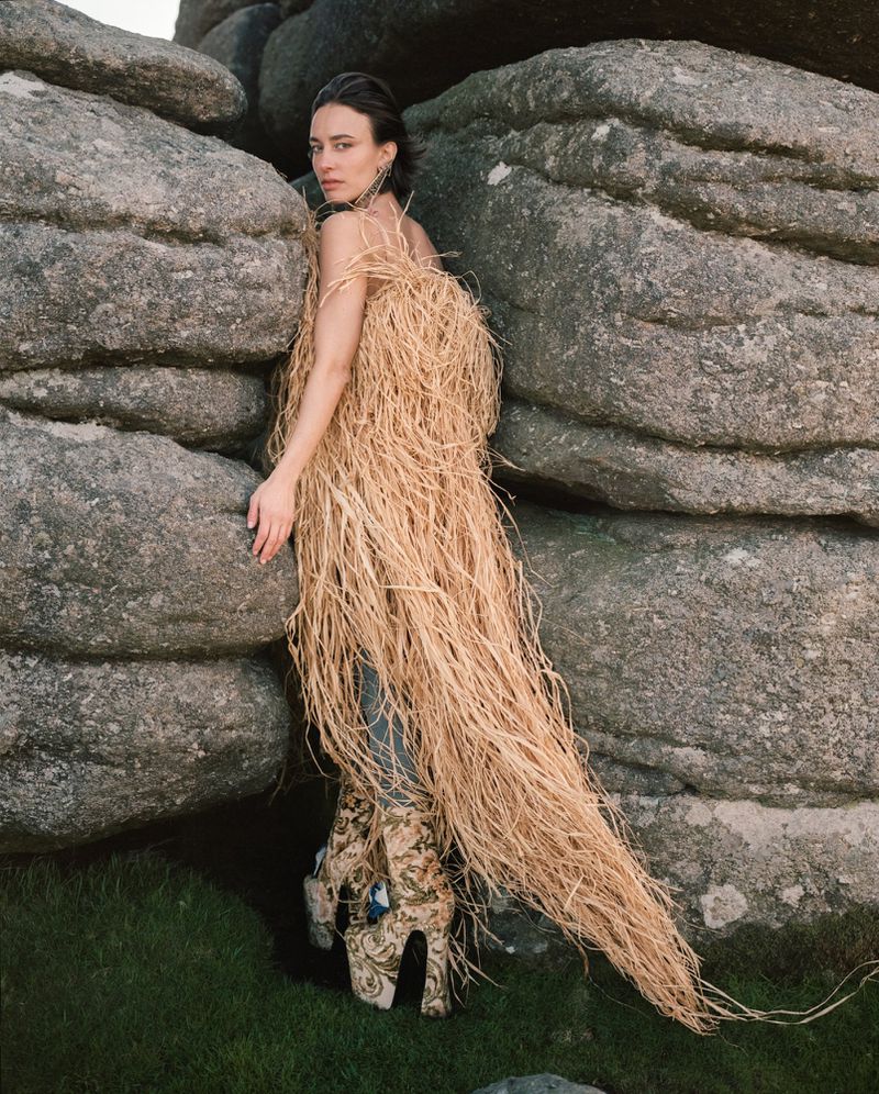 Model wearing a textured straw garment and patterned platform boots standing among large weathered rocks on a moor