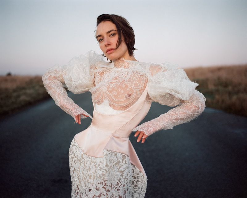 Model wearing a white lace dress with sheer puff sleeves standing on an empty outdoor road under an overcast sky.